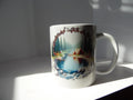 White 11 oz ceramic mug with lakeside camping artwork featuring evergreen trees, blue water and a tent